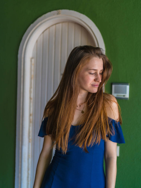 Lifestyle of a young red-haired Caucasian girl in a blue dress in a summer perched next to a white door - Valokuva, kuva