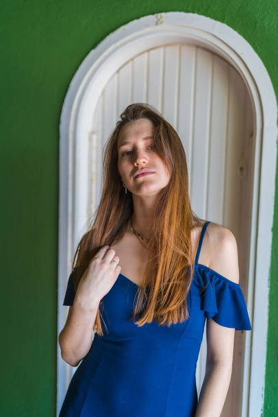 Lifestyle of a young red-haired caucasian girl in a blue dress in a summer perched next to a white door of a green house - Fotografie, Obrázek