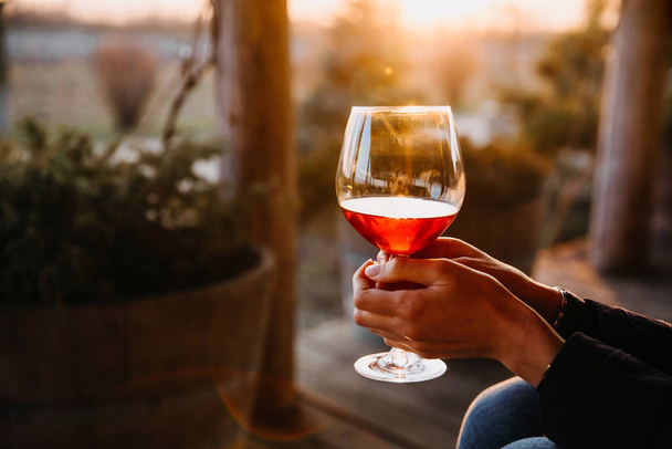 日没の光の中で屋外で赤ワインのガラスを保持する女性の手. - 写真・画像