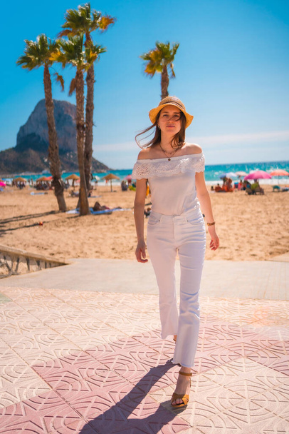 A Caucasian redhead dressed in white and with a straw hat walking along the beach of Calpe in summer, Valencia. Spain - Valokuva, kuva