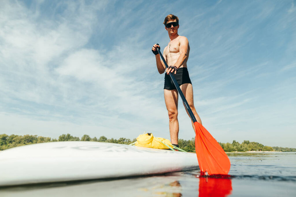 Atletische man met een zonnebril zwemt op een sup board op de vijver, peddelt met een serieus gezicht en kijkt naar de camera. Actieve zomervakantie aan de vijver. - Foto, afbeelding