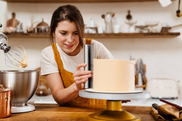 Beautiful pleased pastry chef woman making cake with cream at cozy kitchen - Фото, изображение