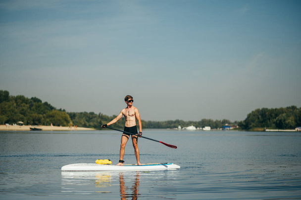 Αθλητικός νεαρός άνδρας κουπιά στέκεται σε μια σανίδα στο ποτάμι και κοιτάζει προς τα πλάγια. Ένας μυώδης άνδρας είναι ενεργός στο νερό.. - Φωτογραφία, εικόνα