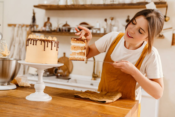 Beautiful happy pastry chef woman showing piece of cake with cream at cozy kitchen - Foto, Imagen