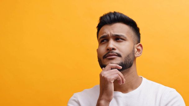 Portrait of young indian man with hand over his beard thinking. Isolated on yellow background - Фото, изображение