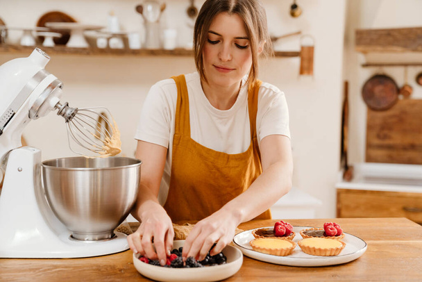 Hermosa mujer pastelera concentrada haciendo tartas con bayas en la acogedora cocina - Foto, imagen