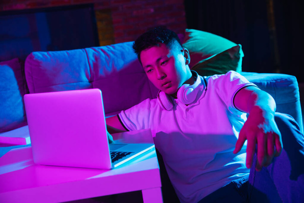 Young Korean man using laptop at home interior in gradient pink blue neon light - Photo, Image