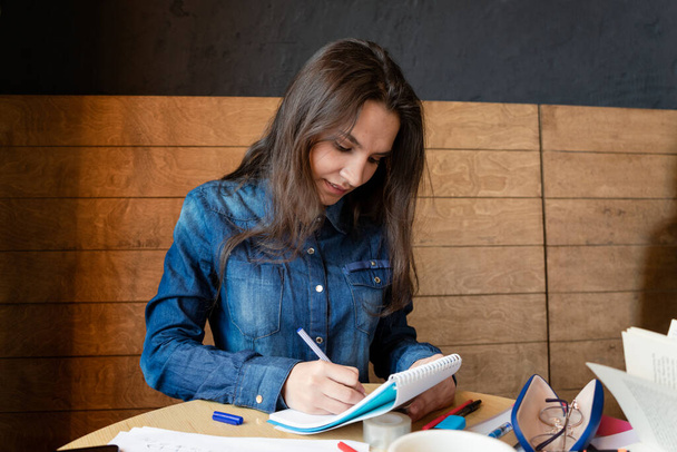 A cheerful girl in a blue denim shirt sitting in a cafe makes notes in a notepad with a pen. Sheets, adhesive tape - Fotografie, Obrázek