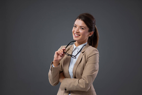 A woman with glasses doing her job. Portrait of a confident adult business woman standing in front of a gray wall holding glasses and touching her chin with glasses. Management, friendly attitude - Foto, afbeelding