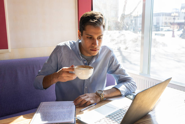 a young businessman looks into a laptop and drinks cappuccino coffee, a notebook with his notes lies next to him, he is in a cafe - Foto, Imagem