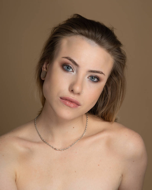 Excite girl with heterochromia, nose piercing and plug in one ear, and strange hairstyle. She has amazing professional makeup and silver chain around her neck. Beige background. Studio shot - Photo, Image