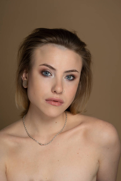 Sexual girl with heterochromia, nose piercing and plug in one ear, and strange hairstyle. She has amazing professional makeup and silver chain around her neck. Beige background. Studio shot - Photo, Image