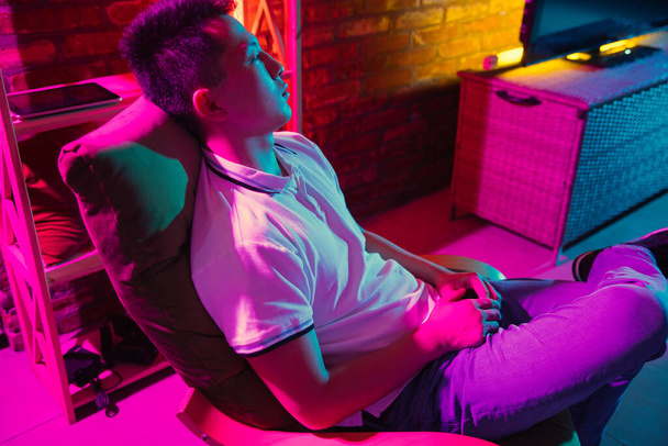 Young Korean man in headphones at home interior in gradient pink neon light - Foto, afbeelding