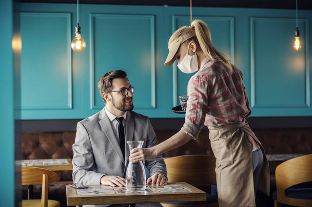 Restaurant service and corona virus. The blonde waitress is wearing a uniform and a protective face mask. She puts a bottle of water on the table and serves the guest in a suit in a modern restaurant - Фото, изображение