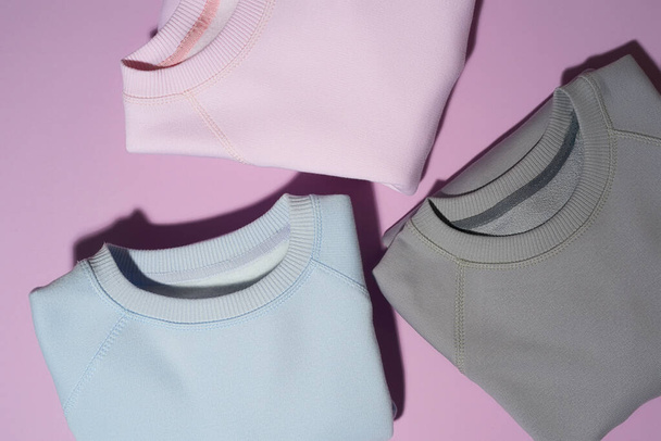 Closeup shot of three folded pastel pink, blue and gray sweatshirts isolated on pale purple background - Foto, imagen