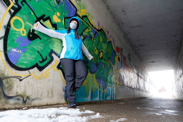 νεαρή γυναίκα σε μπλε άνορακ με κουκούλα και προστατευτική μάσκα ακουμπισμένη σε έναν τοίχο γκράφιτι σε μια υπόγεια διάβαση και με τα χέρια απλωμένα                     - Φωτογραφία, εικόνα