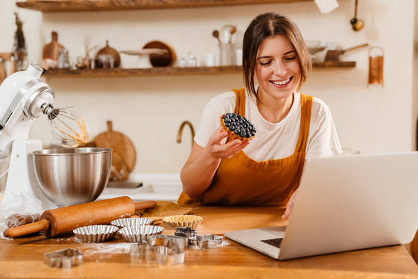 Happy pastry chef woman showing tart while making video call on laptop at cozy kitchen - Фото, зображення