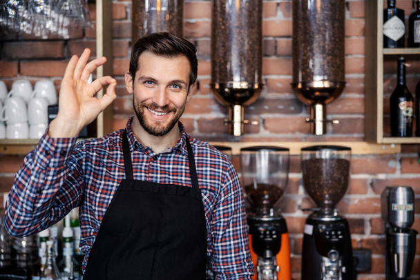 Restoran servisi ve kafe. Yetişkin bir erkek garson bir kafede bir barın arkasında duruyor ve el işareti gösteriyor. Hoş bir gülümseme ve olumlu bir tavır. Kafeterya ve kahve yapma. - Fotoğraf, Görsel