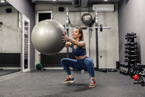 Squat with a big gray ball. An attractive woman in sportswear does exercises to burn the muscles of the buttocks legs and arms. The female crouches and holds a large Pilates ball at shoulder height - Photo, Image