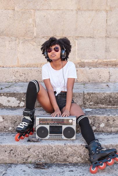 Young black woman with afro hair, roller skates and sunglasses listens to music with headphones from an old radio cassette player while sitting on a stone stairs outdoors on a hot summer day - Foto, Imagem