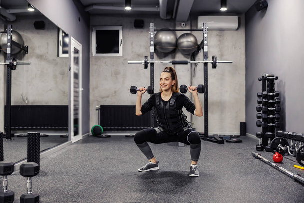 Electrical muscle stimulation. A young and healthy woman in a special black EMS suit performs squats and does arm exercises with dumbbells in a modern gym concept. Revolution in training - Photo, Image