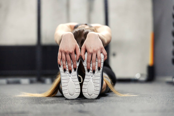 Muscle stretching in spot training. A female person sits on the gym floor and stretches her leg and foot muscles. Women's hands hold the sole of the sneaker and stretch the leg muscles, cool down - Photo, Image