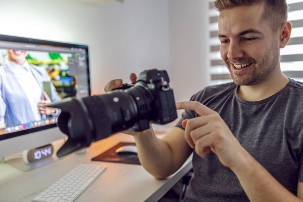 A portrait of a man in an indoor workspace who looks with a smile at the photos he took while filming on the camera he holds in his hands. Freelance job, home office, cozy indoor workspace - Valokuva, kuva