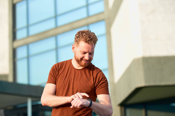 Young sporty ginger man looking at the watch after exercising outdoors. - Φωτογραφία, εικόνα