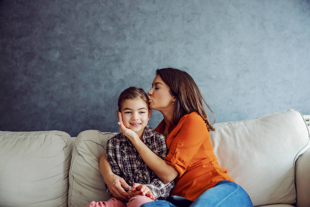 Счастливая мать обнимает и целует свою любимую дочь, сидя на диване. Концепция счастливой семьи. - Фото, изображение