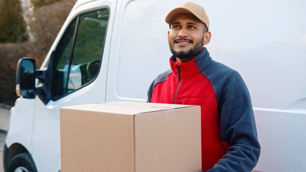 Uomo di consegna sorridente mentre tiene il pacco davanti al furgone di trasporto - Foto, immagini