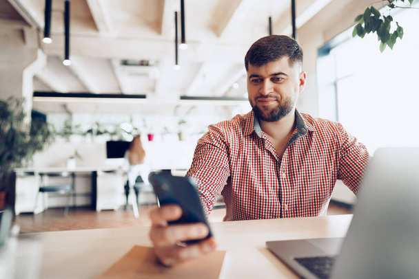 Young man in casual shirt using smartphone in office while sitting at his desk - Foto, Bild