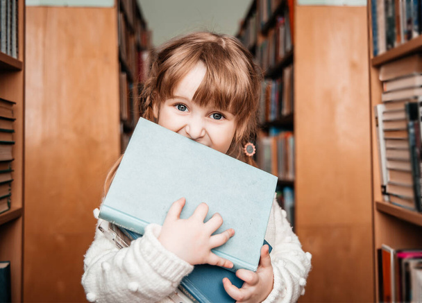 Baby girl in the library with books in her hands. cute toddler explores the bookshelves - Fotoğraf, Görsel