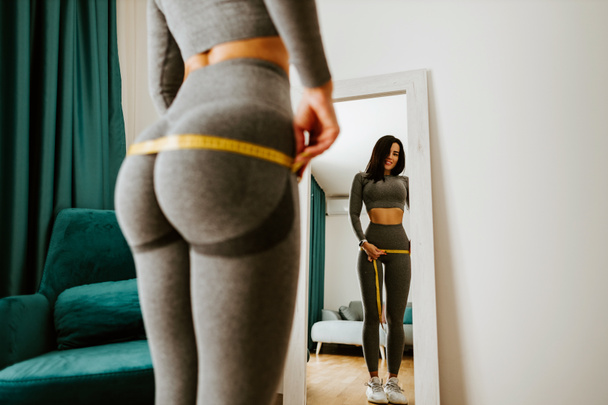 Fitness Woman Measuring body With Tape Standing In Front Of Mirror At Home - Φωτογραφία, εικόνα