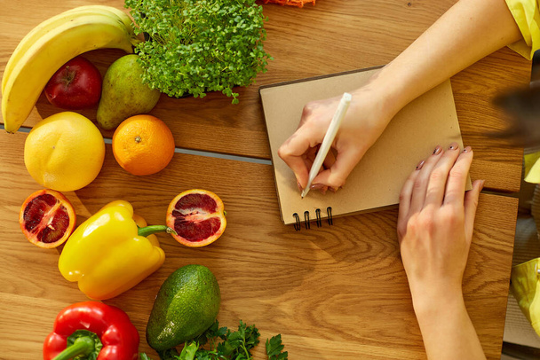 Vrouw van plan, het schrijven van wekelijkse maaltijden op een maaltijd planner nota of dieet plan op houten tafel met gezonde voeding groenten en fruit in haar keuken thuis. - Foto, afbeelding