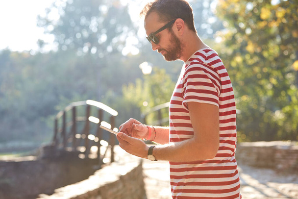 Jonge volwassen man met moderne smartphone in het park. - Foto, afbeelding
