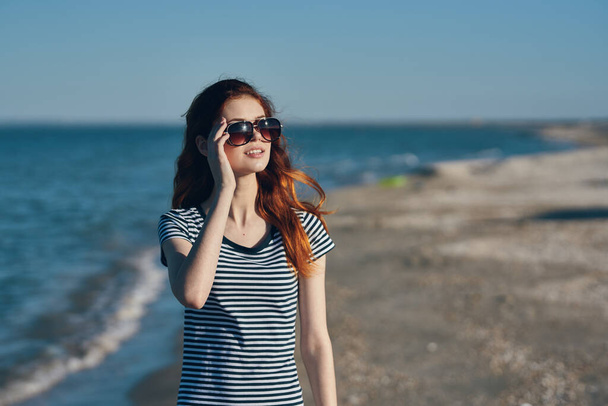 portret van vrouw in t-shirt en bril op het strand in de bergen bij de zee - Foto, afbeelding