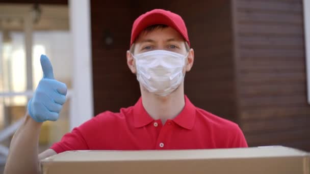 Egy biztonsági maszkot viselő szabadító arcképe, hüvelykujjas dobozzal a kezében. Szállítási szolgáltatás a karanténban coronavirus covid-19. - Felvétel, videó