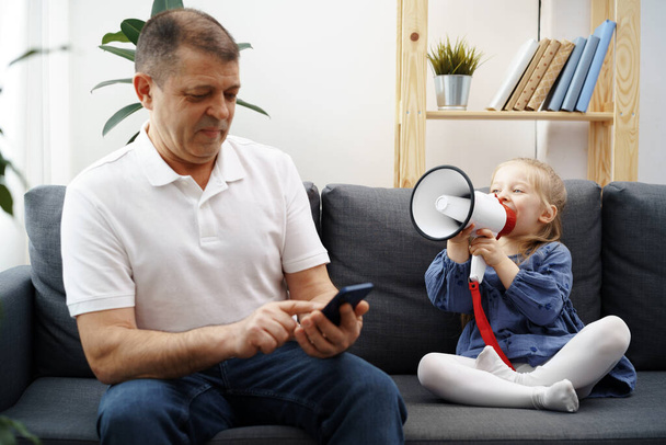 Grandpa sitting on sofa and using smartphone, little granddaughter shouting through megaphone to him to stop - Foto, Imagen