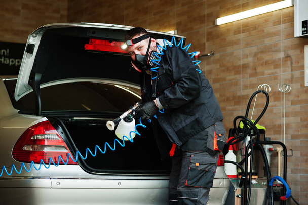 Man in uniform and respirator, worker of car wash center, cleaning car interior with hot steam cleaner. Car detailing concept. - Foto, Bild