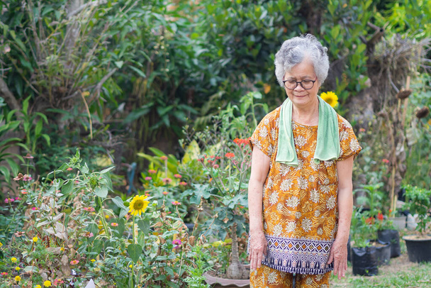 Portrait of an elderly woman exercises, smiling and looking down while standing in a garden. Space for text. Concept of aged people and healthcare. - Photo, Image