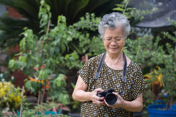 Femme âgée avec les cheveux gris courts, souriant et tenant l'appareil photo numérique tout en se tenant dans un jardin. Espace pour le texte. Concept de personnes âgées et photographie. - Photo, image