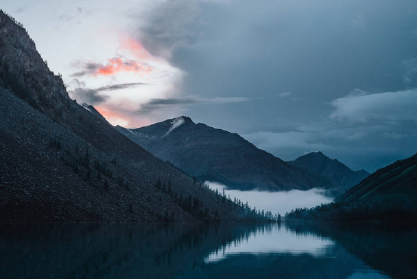 Low cloud above highland lake. Silhouettes of trees on hillside along mountain lake in dense fog. Reflex of pines to calm water. Alpine tranquil landscape at early morning. Ghostly atmospheric scenery - Photo, Image