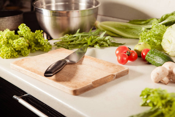 Cucinare l'insalata in cucina. Sul piano di lavoro c'è un tagliere e un coltello. Verdure ed erbe sono disposte nelle vicinanze - Foto, immagini