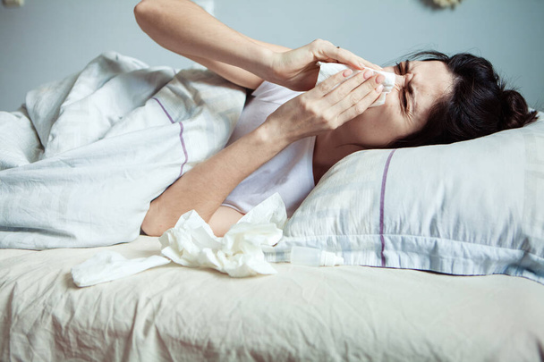 Μια νεαρή γυναίκα αισθάνεται κρύο σε ένα κρεβάτι. Προσπαθεί να ζεσταθεί κάτω από την κουβέρτα. Το άρρωστο κορίτσι θάβει μια σταγόνα κρύου στη μύτη. Έννοια κρύου καιρού. Γυναίκα ξαπλωμένη στο κρεβάτι και με πυρετό  - Φωτογραφία, εικόνα