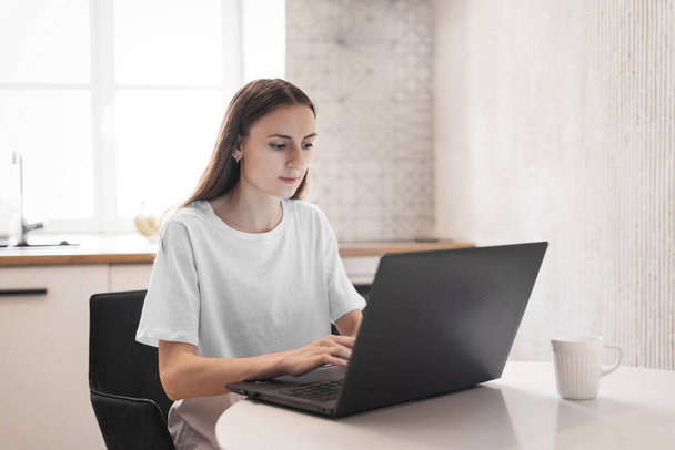 Mladá žena pracuje na laptopu v kuchyni. Žena na volné noze pracuje online doma - Fotografie, Obrázek