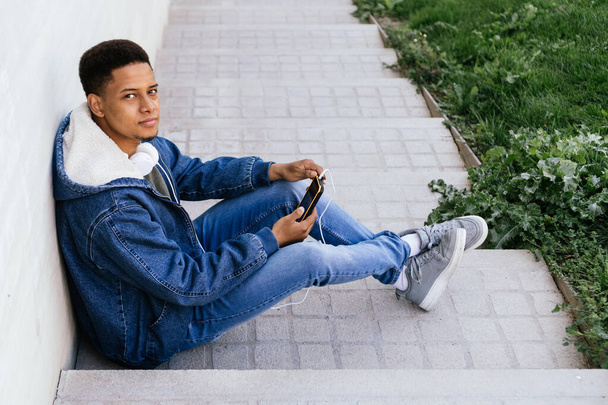 Πορτρέτο ενός Λατίνου που χρησιμοποιεί το κινητό του τηλέφωνο και τα ακουστικά του στο δρόμο. Νεαρός Αφρο-αγόρι χρησιμοποιεί ένα κινητό τηλέφωνο σε έναν απλό γκρίζο τοίχο. Οριζόντια φωτογραφία με χώρο αντιγραφής  - Φωτογραφία, εικόνα