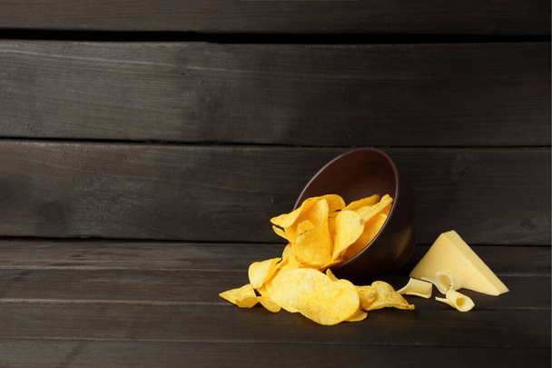 Kartoffelchips in einer Tonschale. Kopfüber Teller mit Kartoffelchips, ein Stück Käse auf dunklem Holzhintergrund mit Kopierraum. Banner. - Foto, Bild