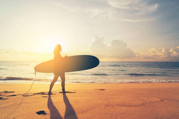 Frau hält Surfbrett am tropischen Sonnenuntergang Strand Hintergrund. Sommerferien- und Sporterlebniskonzept. Vintage Ton Filtereffekt Farbstil. - Foto, Bild