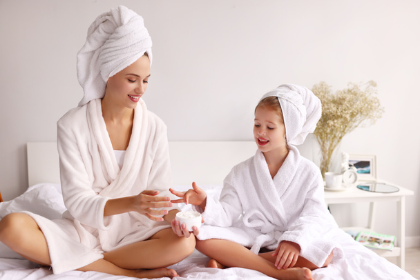 Cheerful mother and daughter smiling   while sitting on bed and smearing cream on face during skin care routine at home - Photo, Image