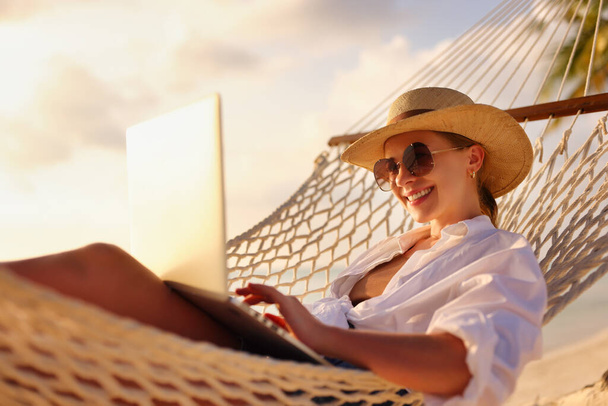 遠隔操作だ。ビーチのハンモックで日没時にリラックスしながら、ラップトップで働く若い幸せな女性、わらの帽子とサングラスの女性のフリーランスの側面図。休暇中の距離仕事 - 写真・画像